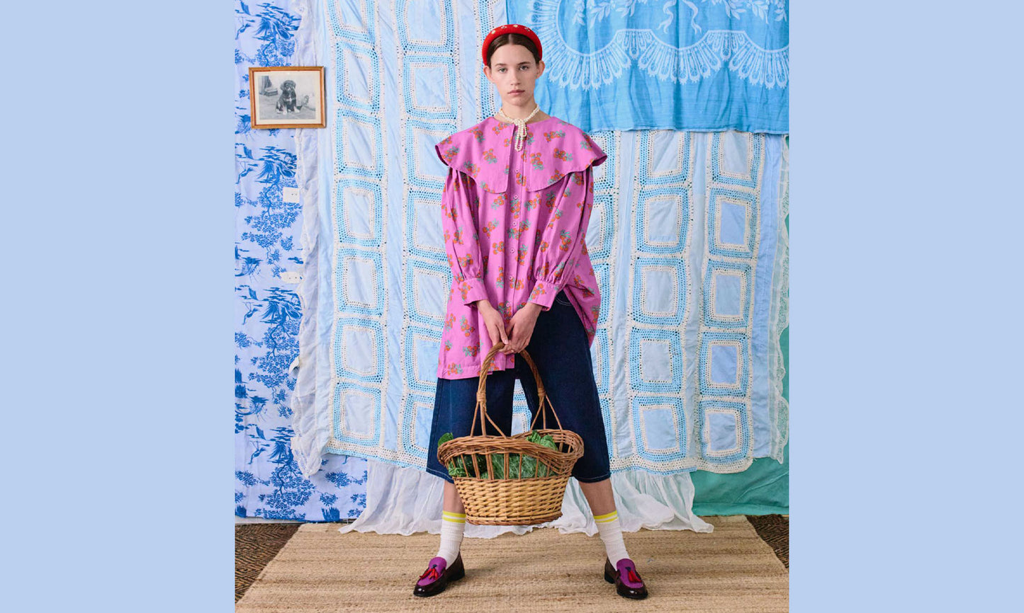 Person wearing a pink patterned shirt and blue jeans holding a woven basket in a room with blue patterned curtains.